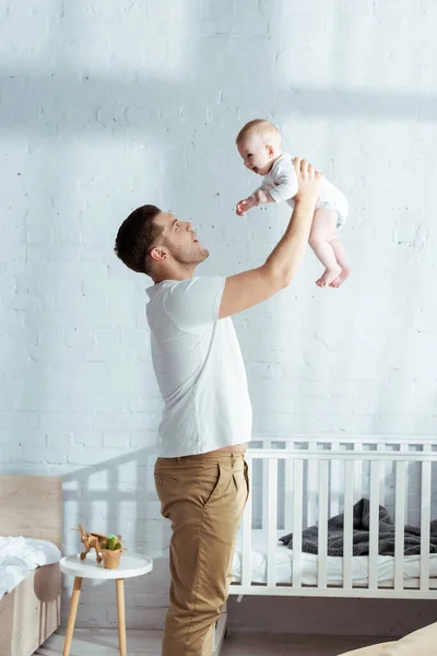 Happy father holding adorable baby boy in outstretched hands near baby cot — Stock Photo