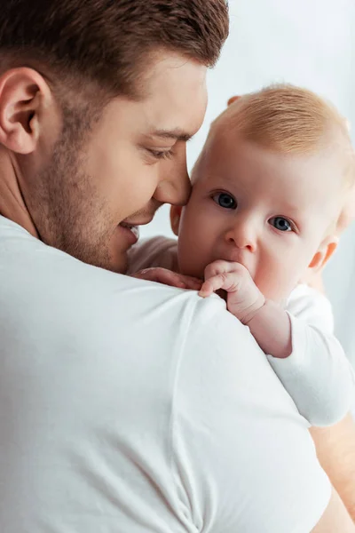 Happy father holding adorable baby boy looking at camera with hand near mouth — Stock Photo