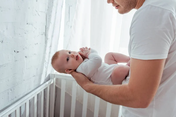 Обрезанный вид отца, держащего очаровательного мальчика над детской кроваткой — стоковое фото