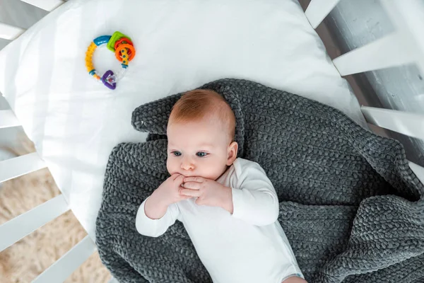 Top view of adorable baby boy lying in baby cot on blanket with hands near mouth — Stock Photo