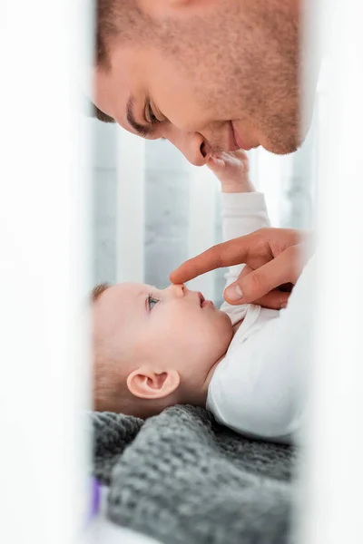 Enfoque selectivo feliz joven padre tocando la nariz de adorable pequeño hijo acostado en cuna - foto de stock
