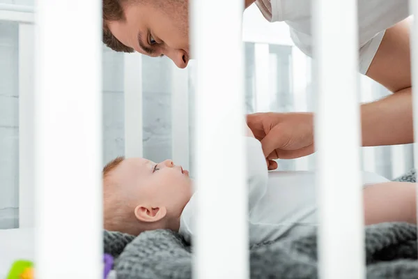 Foyer sélectif de jeune homme penché au-dessus adorable petit fils couché dans le lit de bébé — Photo de stock