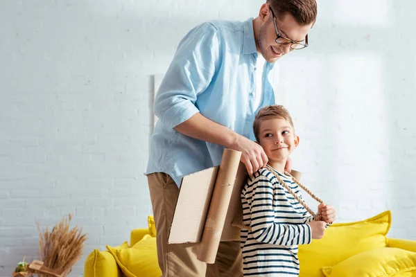 Sonriente padre fijación de cartón plano alas en la parte posterior de adorable hijo — Stock Photo