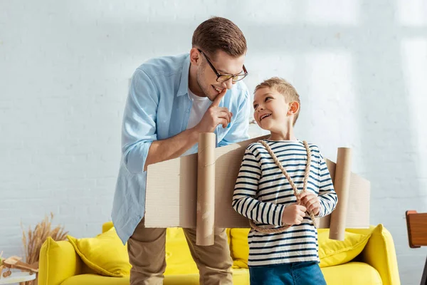 Cheerful father showing hush gesture to happy son with cardboard wings behind back — Stock Photo