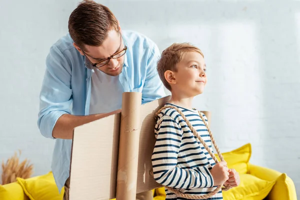 Concentrated father fixing carton plane wings on back of happy son — Stock Photo