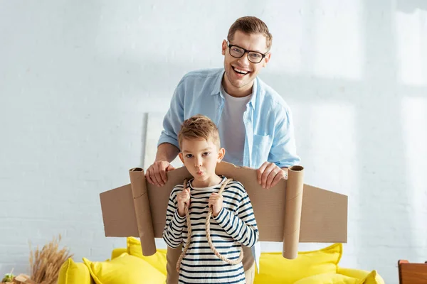 Glücklicher Vater und entzückender Junge mit Kartonflügeln auf dem Rücken, der in die Kamera blickt — Stockfoto