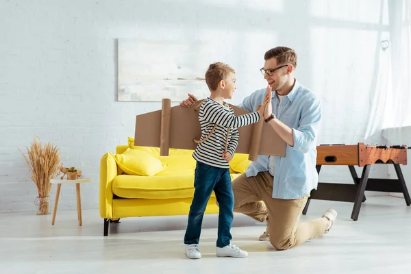 Happy boy with carton plane wings on back giving high five to smiling father — Stock Photo