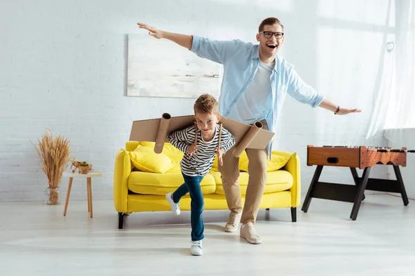 Happy boy with carton plane wings, and cheerful father having fun while imitating flying — Stock Photo