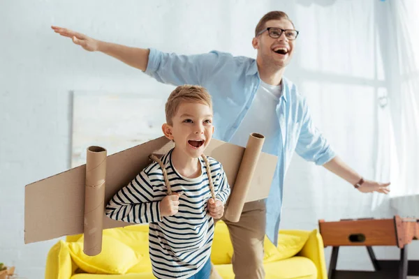 Niño feliz con alas de avión de cartón, y padre alegre que se divierte mientras imita volar - foto de stock