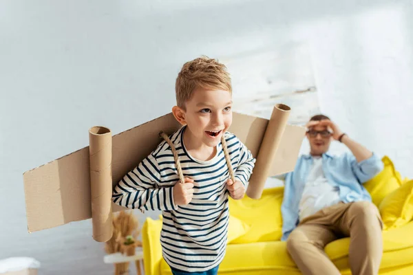 Enfoque selectivo de niño feliz con alas de avión de cartón en la espalda imitando volar cerca de padre - foto de stock