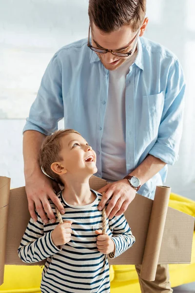 Lächelnder Vater berührt Schultern eines glücklichen Sohnes mit Flugzeugflügeln aus Pappe auf dem Rücken — Stockfoto