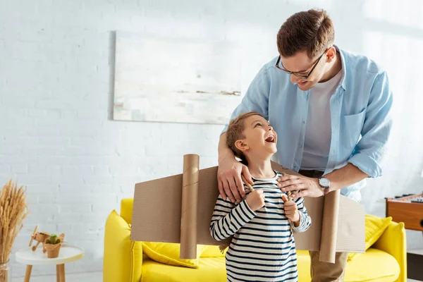 Smiling father touching shoulders of adorable son with cardboard plane wings on back — Stock Photo