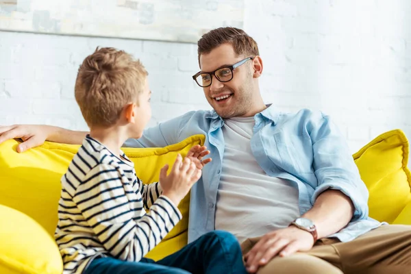 Lächelnder Mann im Gespräch mit entzückendem Sohn, während er zu Hause auf dem Sofa sitzt — Stockfoto