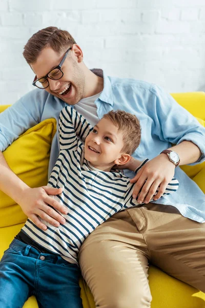 Feliz padre e hijo riendo mientras están sentados juntos en casa - foto de stock