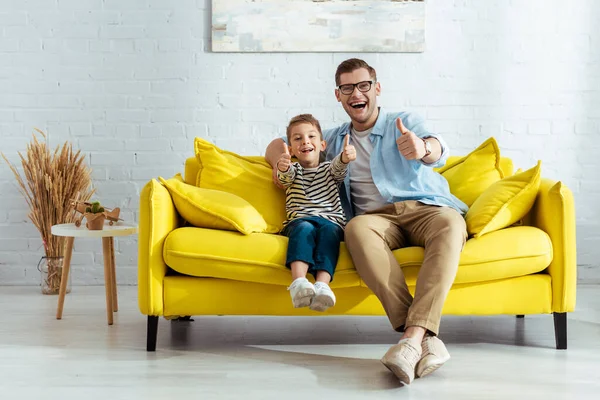 Збуджений батько і син показують великі пальці вгору, сидячи на жовтому дивані — стокове фото