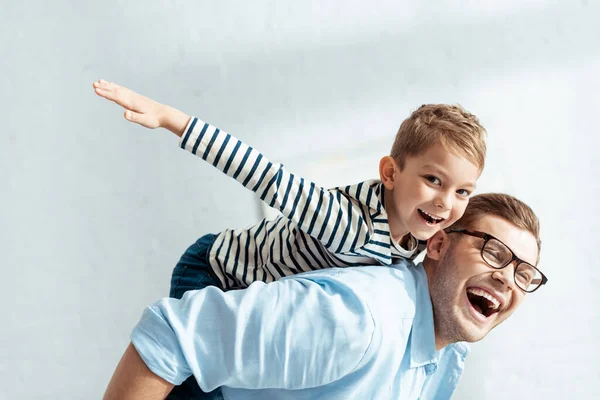 Fröhlicher Vater huckepack glücklicher Sohn blickt in die Kamera und imitiert das Fliegen — Stockfoto