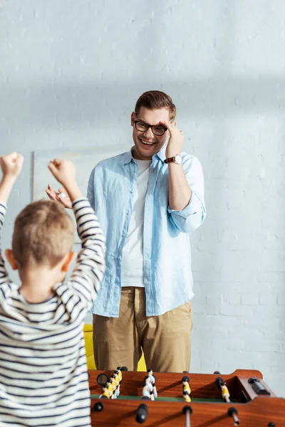 Vista posterior del niño emocionado mostrando el gesto ganador mientras juega al futbolín con el padre - foto de stock