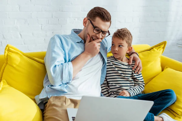 Joven padre riendo y abrazando hijo mientras está sentado en el sofá y el uso de la computadora portátil - foto de stock