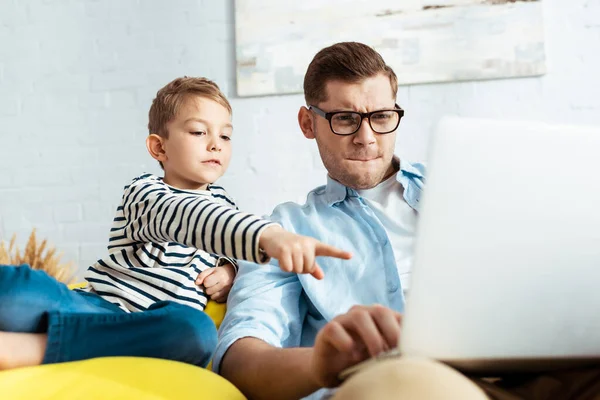 Прелестный мальчик, указывающий пальцем на ноутбук рядом с сконцентрированным отцом — стоковое фото