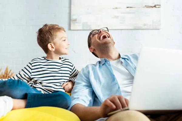 Menino sorrindo olhando para o pai feliz usando laptop — Fotografia de Stock