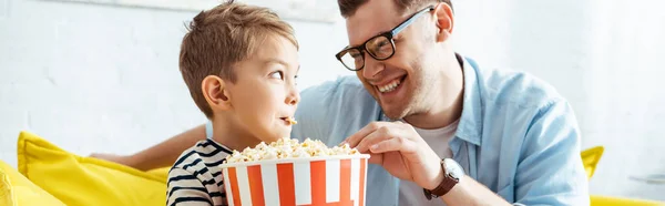Horizontal image of happy father and son looking at each other while eating popcorn from bucket — Stock Photo
