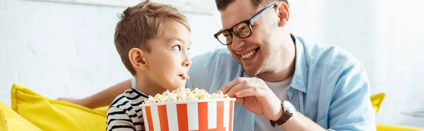 Imagem horizontal de pai e filho felizes olhando um para o outro enquanto comem pipocas de balde — Fotografia de Stock