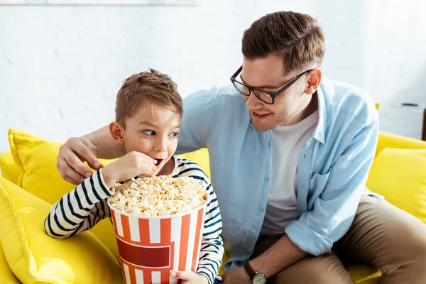 Feliz padre mirando hijo comer palomitas de maíz de cubo - foto de stock