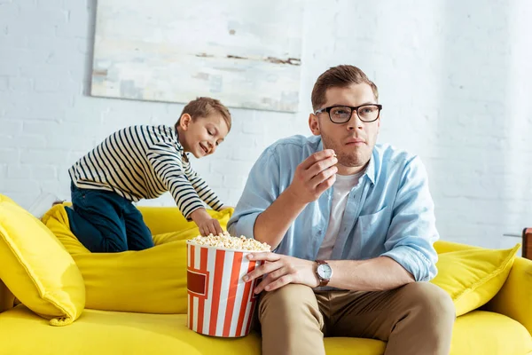 Smiling boy taking popcorn from bucket while attentive father watching tv — Stock Photo