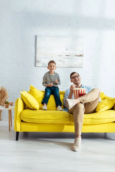 Happy father and son sitting on yellow sofa, watching tv and eating popcorn — Stock Photo