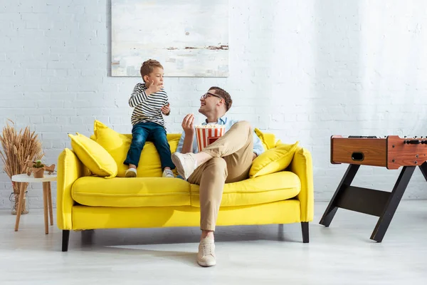 Felice padre e figlio mangiare popcorn mentre seduto sul divano giallo — Foto stock