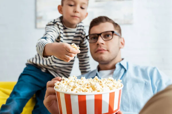 Selektiver Fokus des niedlichen Jungen, der Popcorn aus dem Eimer nimmt, während der konzentrierte Vater fernsieht — Stockfoto