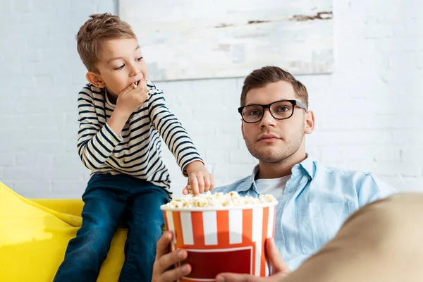 Lindo chico tomando palomitas de maíz de cubo mientras concentrado padre viendo tv - foto de stock