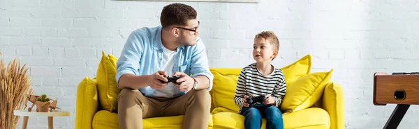KYIV, UKRAINE - JUNE 9, 2020: horizontal image of young father and cute son sitting on yellow sofa with joysticks — Stock Photo