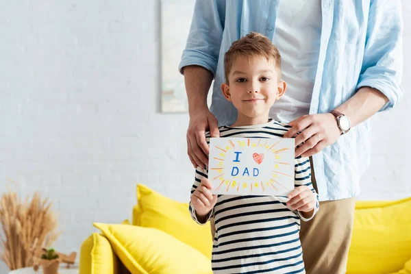 Vista recortada del hombre tocando los hombros del hijo sosteniendo la tarjeta de felicitación del día de los padres hechos a mano con letras y símbolo del corazón - foto de stock