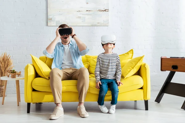 Excited father and son using vr headsets while sitting on yellow sofa — Stock Photo