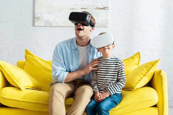 Shocked man and son using vr headsets while sitting on yellow sofa — Stock Photo