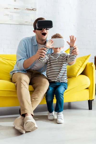 Aufgeregter Mann hält Hände seines Sohnes, während er VR-Headsets zusammen benutzt — Stockfoto