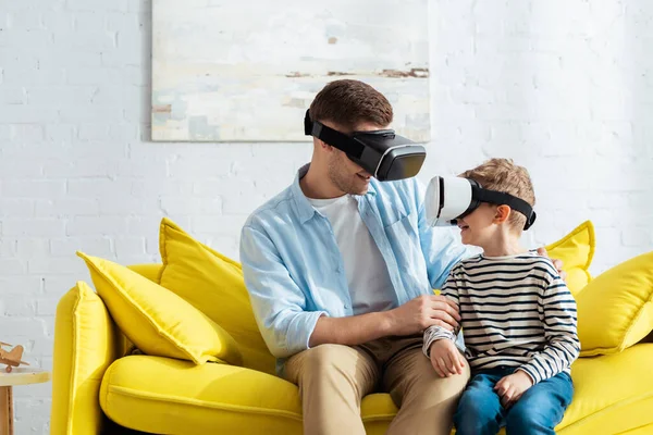 Smiling father and son looking at each other while sitting on sofa in vr headsets — Stock Photo