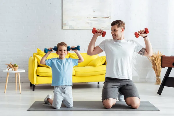 Feliz padre e hijo haciendo ejercicio con pesas mientras están de pie de rodillas en la colchoneta de fitness - foto de stock