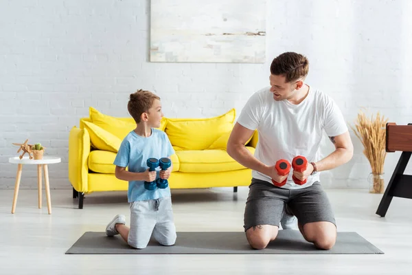 Jeune père et fils regardant l'autre tout en se tenant sur les genoux sur tapis de fitness — Photo de stock