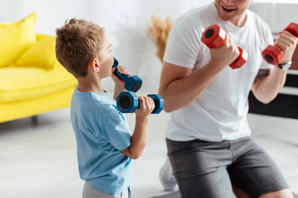 Vista recortada del hombre cerca de hijo haciendo ejercicio con pesas en casa - foto de stock