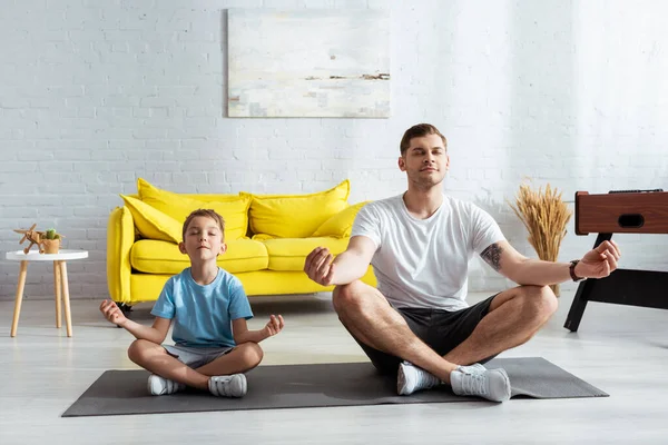Pai e filho sentado em lótus posa no tapete de fitness com os olhos fechados — Fotografia de Stock