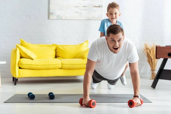 Junger Mann macht Liegestütze mit Hanteln, während Sohn auf dem Rücken sitzt — Stockfoto