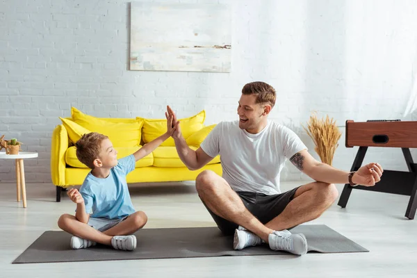 Joyeux père et fils donnant haute cinq tout en étant assis sur tapis de remise en forme dans des poses de lotus — Photo de stock