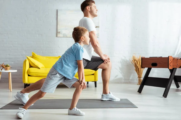 Vista lateral do pai jovem e filho bonito fazendo lunges enquanto se exercita em casa — Fotografia de Stock