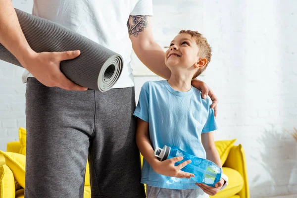 Vista ritagliata dell'uomo che tiene il tappeto di idoneità mentre tocca la spalla del figlio felice che tiene la bottiglia di sport — Foto stock