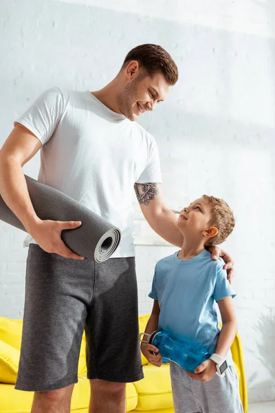 Homem alegre com tapete fitness tocando ombro de filho adorável segurando garrafa de esportes — Fotografia de Stock