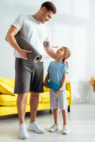 Hombre feliz con tapete de fitness tocando hombro de adorable hijo sosteniendo botella deportiva - foto de stock