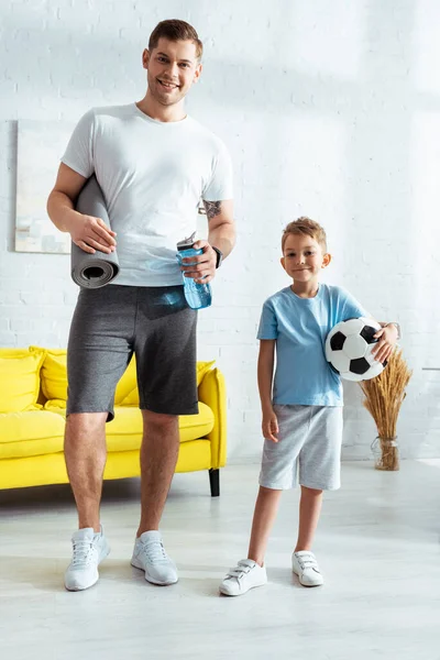 Hombre feliz con tapete de fitness y botella de deportes cerca de hijo sosteniendo pelota de fútbol - foto de stock