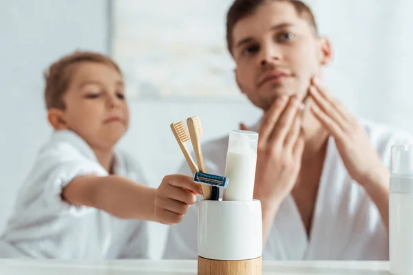 Enfoque selectivo del hombre tocando la cara mientras que el hijo adorable toma cepillo de dientes - foto de stock